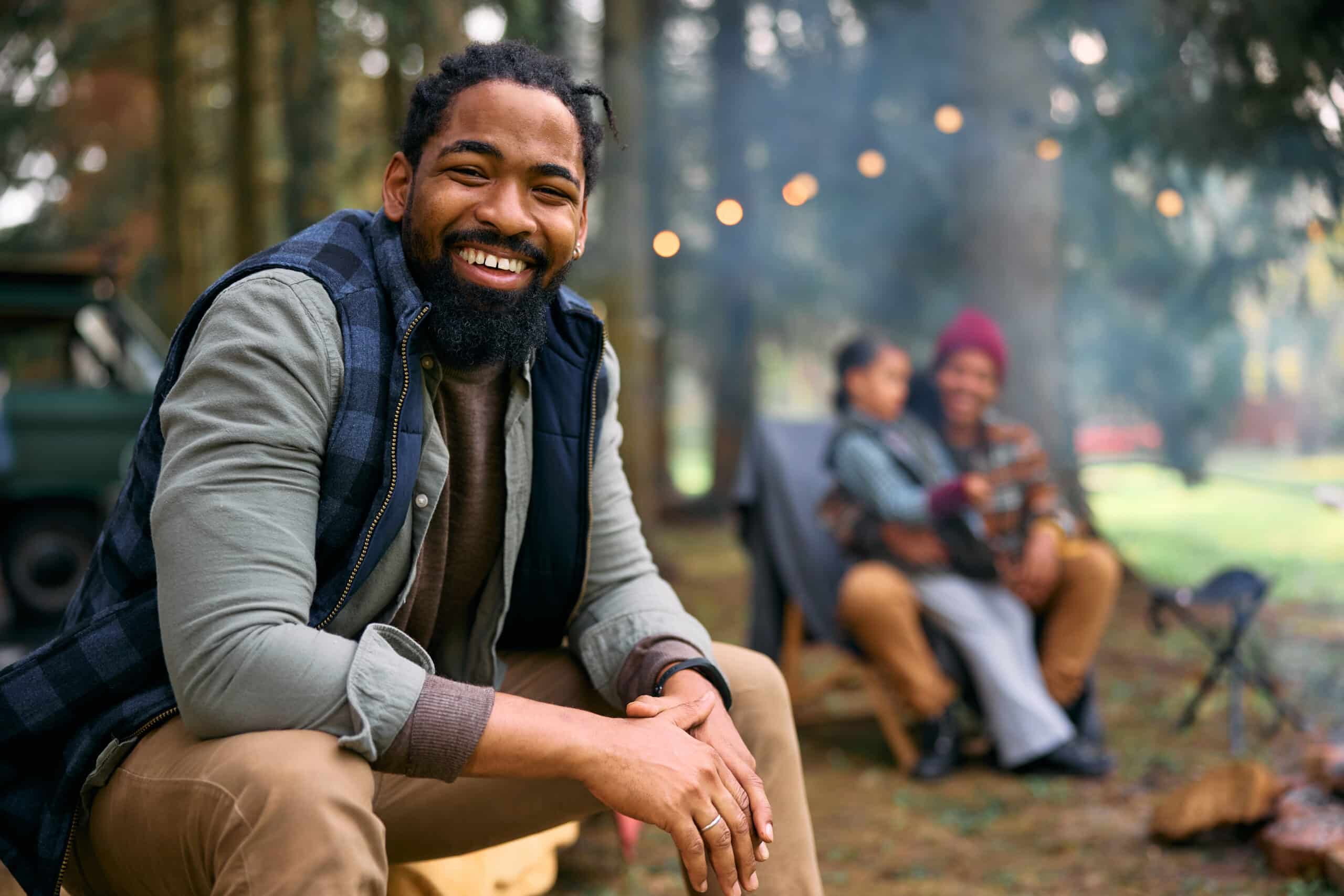 Happy African American father enjoying in camping at trailer park and looking at camera. His family is in the background.