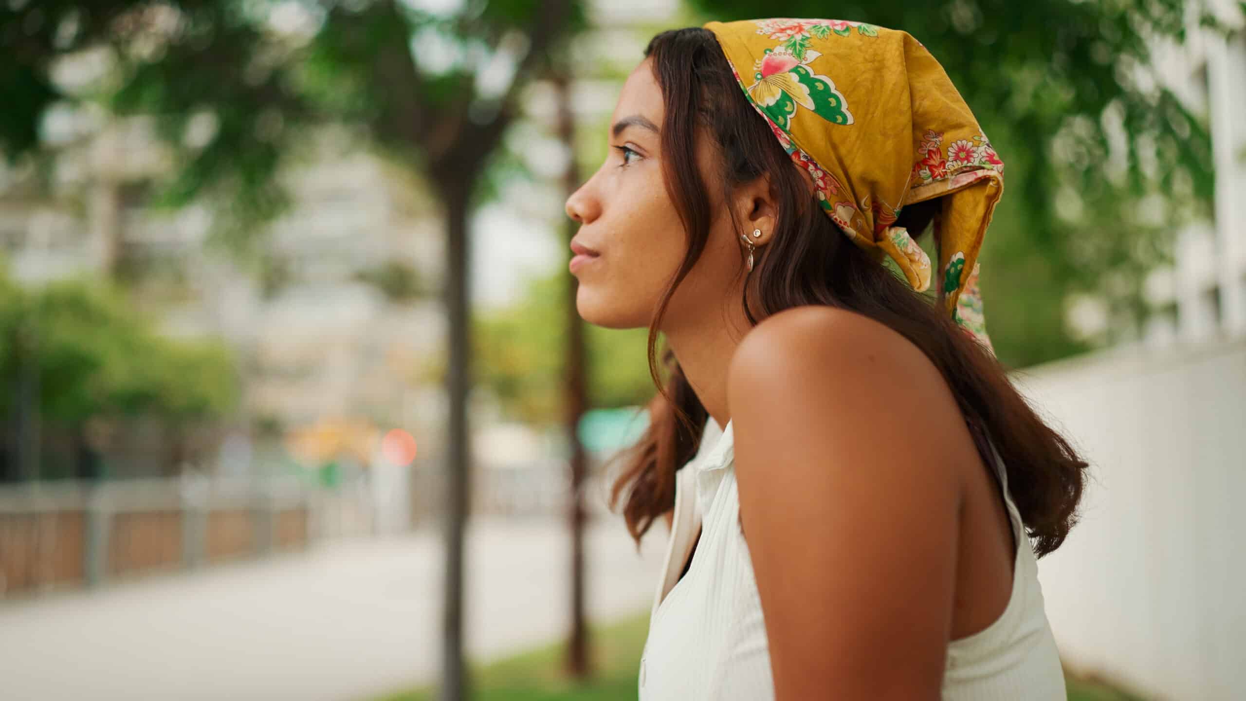 Smiling cute tanned woman with long brown hair wearing white top and yellow bandana seated on bench on cityscape background - self-compassion