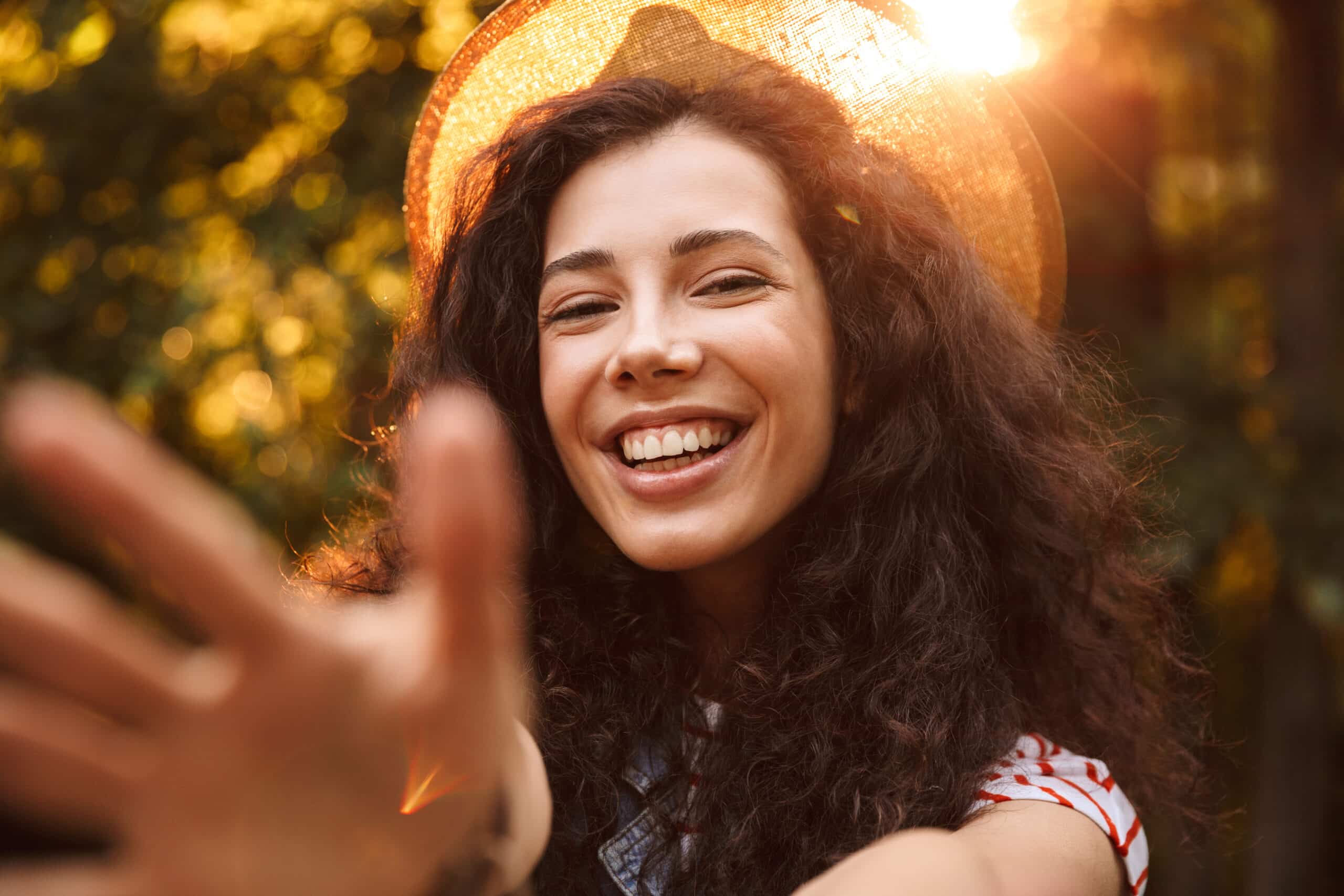 Photo closeup of brunette attractive woman 18-20 wearing summer straw hat smiling and spreading hands to camera while walking through park in sunny day with trees background