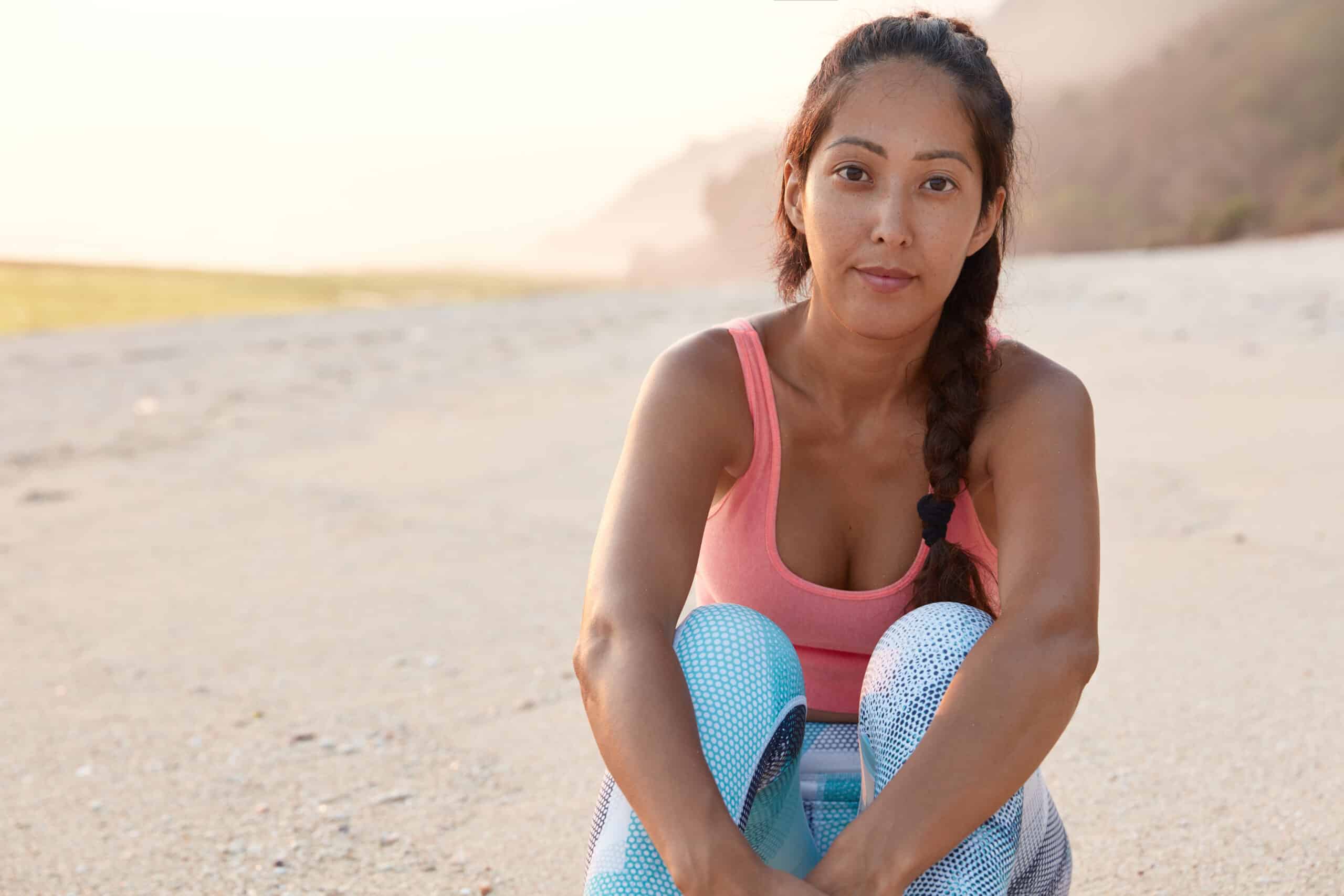 Pleasant looking serious athletic woman has rest after tired training, sits on beach against beautiful sunset with copy space aside for your promotional content. People, sport, motivation, summer